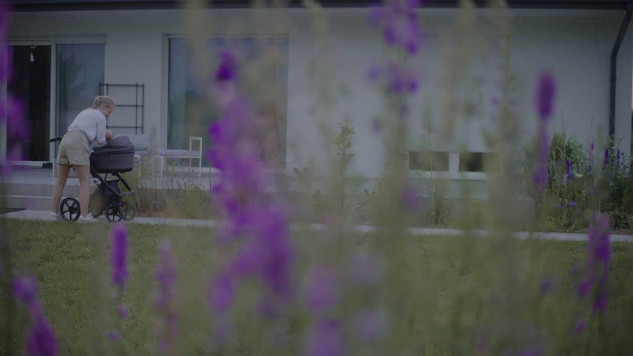 Young Mother Puts Baby in Stroller Near Home and Garden