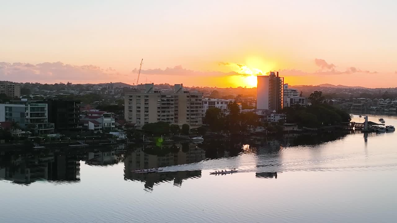 Tre imbarcazioni a quattro con timoniere gareggiano risalendo un tranquillo fiume Brisbane mentre il sole sorge in lontananza.