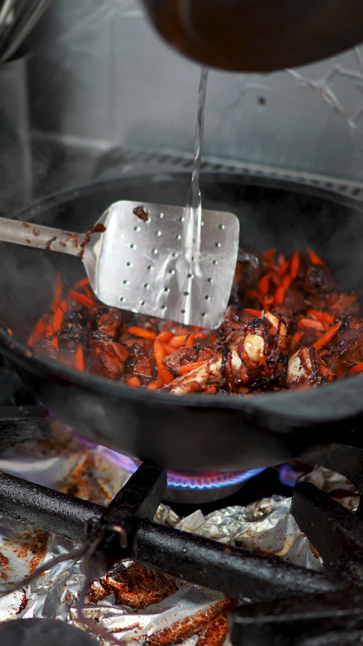 Professional chef pouring hot water from metal pot onto meat, carrots, and onions, carefully mixing ingredients with slotted spoon in kitchen cooking process
