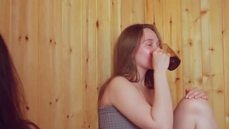 close up of woman fanning herself using sauna hat while two other women relax and raise mugs in toast, all seated in wooden steam room enjoying warmth, comfort, bonding, and shared wellness ritual