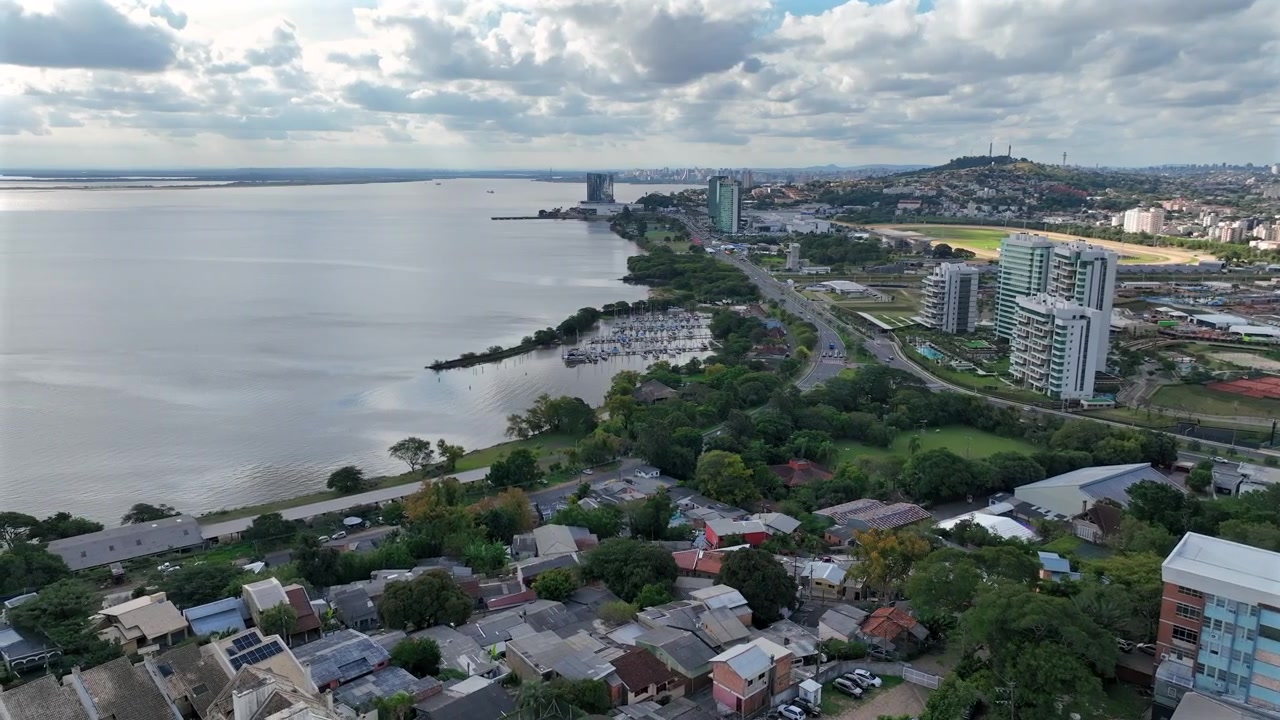 Ilmakuva Porto Alegren eteläisestä järvenrannasta Av. Diário de Notícias -kadun varrella, jossa näkyy venesatama, rakennuksia ja vihreitä kukkuloita.
