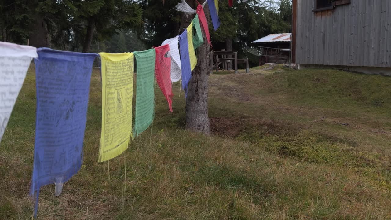 Tibetan prayer flags between trees near a rustic cabin in a calm natural setting. The colorful fabric moves gently in the breeze in daylight. Ideal for travel, culture, or spirituality themes