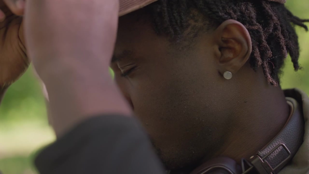 Close up of man adjusting his brown cap in outdoor setting with greenery. subject has dark skin braided hair and is wearing ear gauges soft natural light illuminates scene with blurred