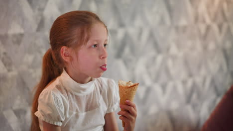 young child with long brown hair wearing white blouse enjoys eating cone of ice cream indoors, focused expression, sitting against artistic patterned wall