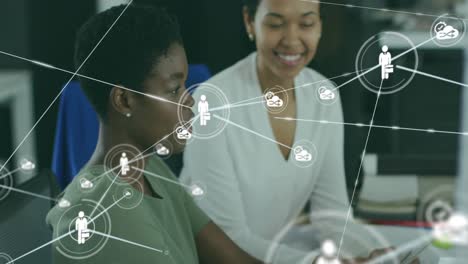 Animation-of-connected-icons-over-diverse-female-coworkers-discussing-reports-on-desktop-at-office