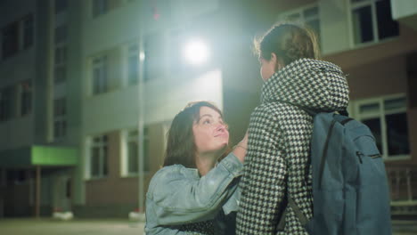 Woman adjusts young girl&#039;s coat as they share caring eye contact outside school entrance early in morning, capturing emotional connection, protective gesture, and warmth in dim outdoor lighting
