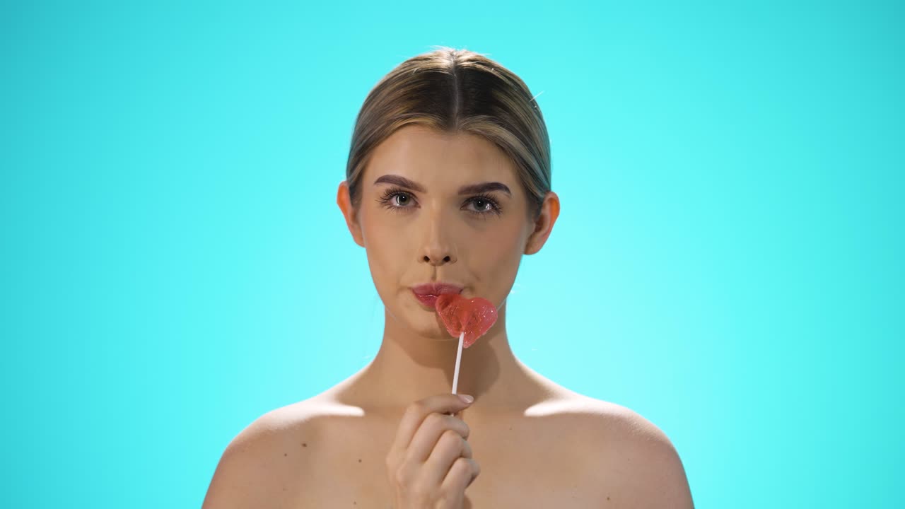 Slow motion Static shot of a young caucasian naked woman licking a heart shaped lollipop while looking sexy into the camera in slow motion against turquoise background