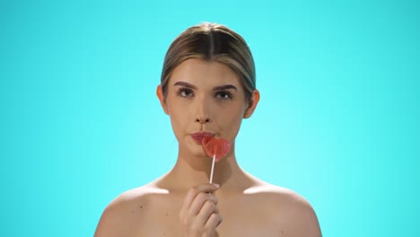 slow motion static shot of a young caucasian naked woman licking a heart shaped lollipop while looking sexy into the camera in slow motion against turquoise background