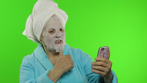 abuela en bata de baño, máscara facial. anciana usando teléfono móvil para llamada de video