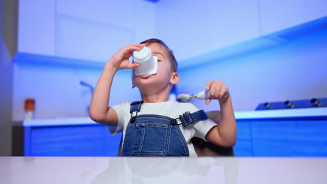 lovely toddler eating yogurt with a spoon. kid takes a cup to his mouth and smudges his face. low angle view.