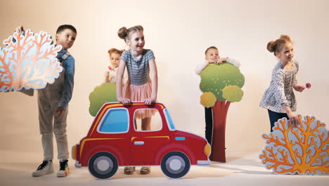 funny children are running with different decorations in the form of cars, trees and bushes on a studio background