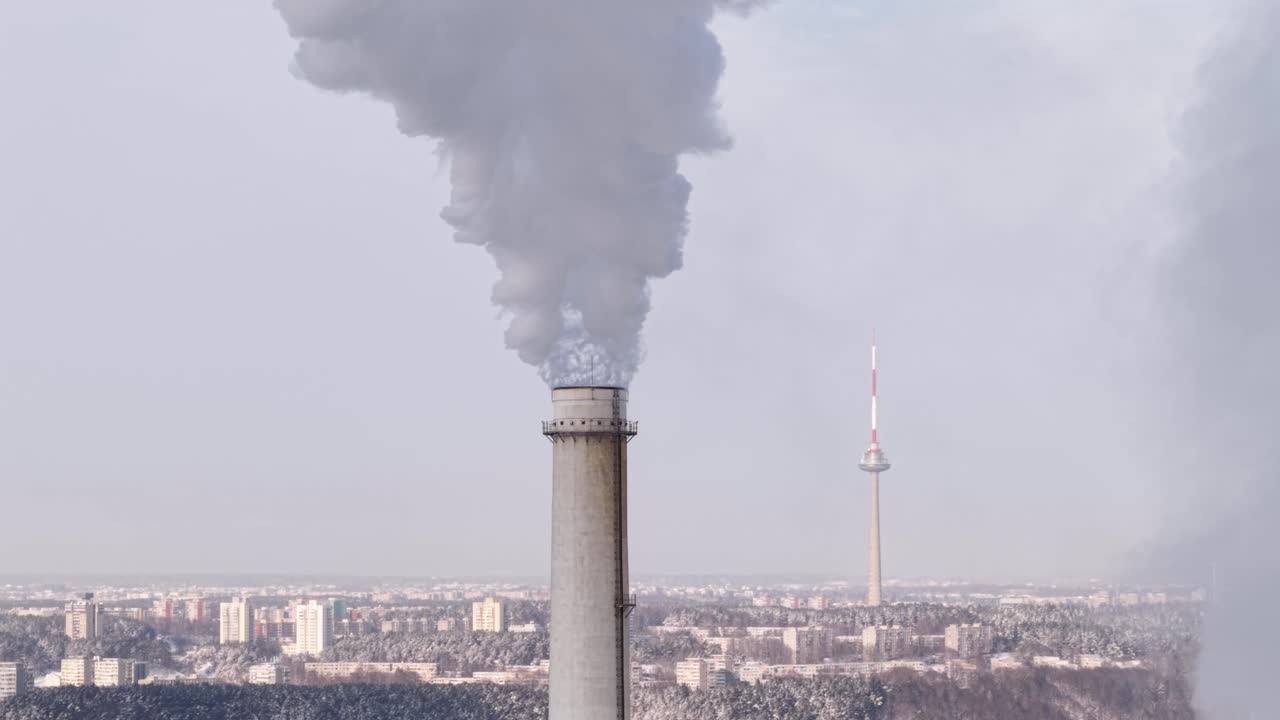 Vista aerea di Vilnius CHP con un fumaiolo che rilascia fumo scuro sopra lo skyline della città e una torre TV in lontananza, che evidenzia l'infrastruttura di riscaldamento urbano e l'inquinamento