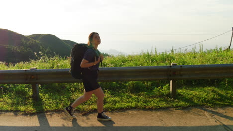 side view tracking woman trekking with large backpack hiking along path in countryside overlooking valley at sunset