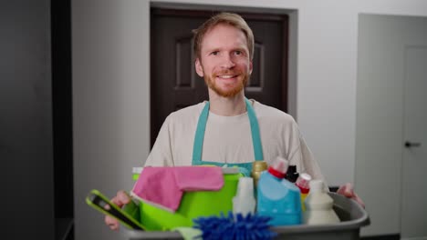 retrato de un hombre de limpieza profesional rubio confiado en un delantal azul que sostiene un cuenco de plástico gris en sus manos que está lleno de una gran cantidad de herramientas de limpia mientras llama a una compañía de limpiezas a la casa en un apartamento moderno