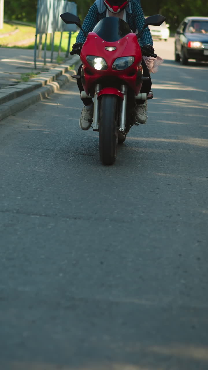 dos parientes viajan juntos en una bicicleta eléctrica roja, ambos con cascos, viajando cerca de una pasarela, con los faros de los coches encendidos, incluido un camión, se ven siguiéndolos en la carretera urbana