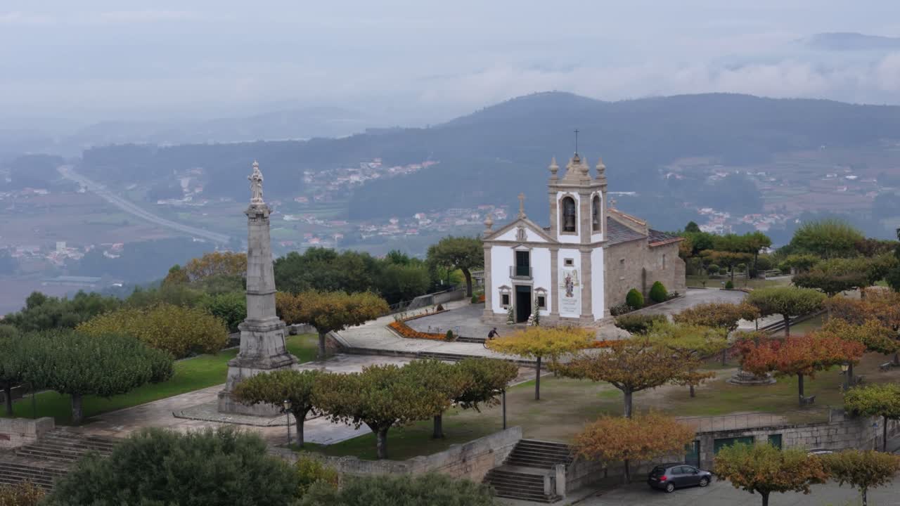 Veduta aerea da drone di una cappella barocca bianca con campanile e colonna di pietra su una collina boscosa.
