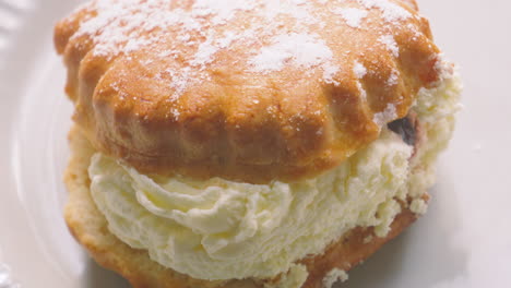 overhead view of delicious traditional cream and jam scone with light dusting of icing sugar on top resting on plate in natural light. british dessert from devon and cornwall. classic afternoon tea.
