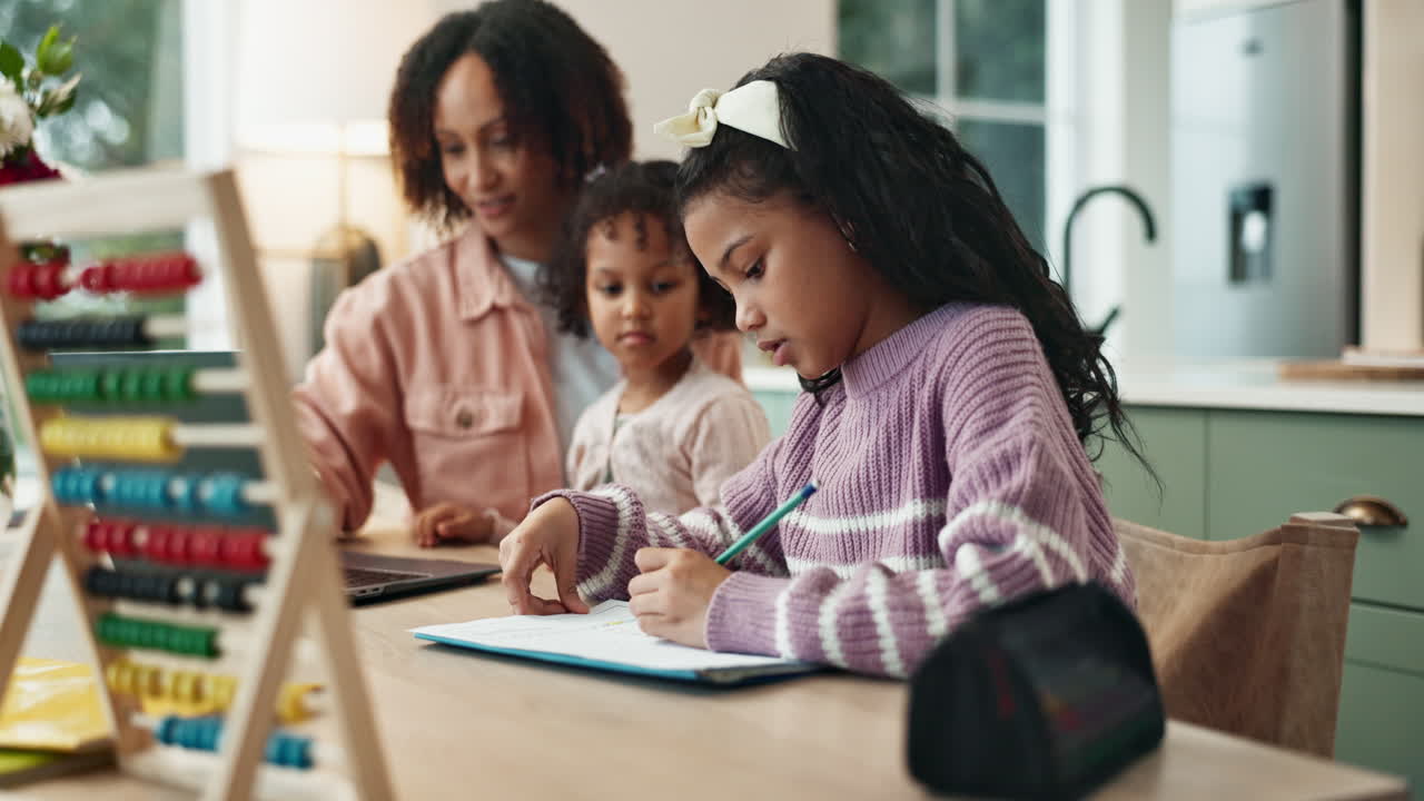 una niña hace su tarea en la mesa de la cocina mientras su madre trabaja en una computadora portátil, con un hermano pequeño sentado a su lado