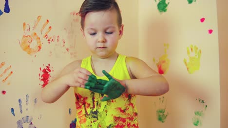 close up of little cute child with painted in green hands making colorful handprints on the wall behind him. playing and having fun.