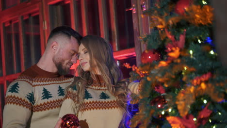 young couple in love hangs christmas balls. happy couple in knitted christmas sweaters decorating christmas tree in their home. christmas tree decoration.