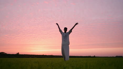 person silhouetted with raised arms in a field at sunset
