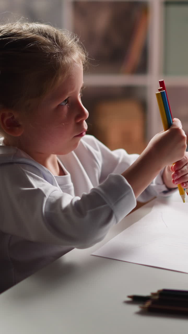 Inspired little artist in white shirt gathers different crayons in bunch to draw on paper sheet sitting at table in light children room slow motion
