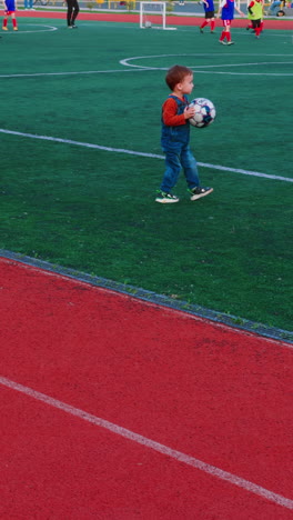 little baby boy outdoor playing with ball. small boy playing football at stadium. vertical video.