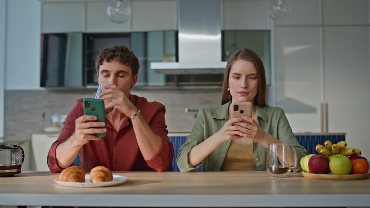 Addicted couple looking mobile phones sitting at kitchen table together closeup