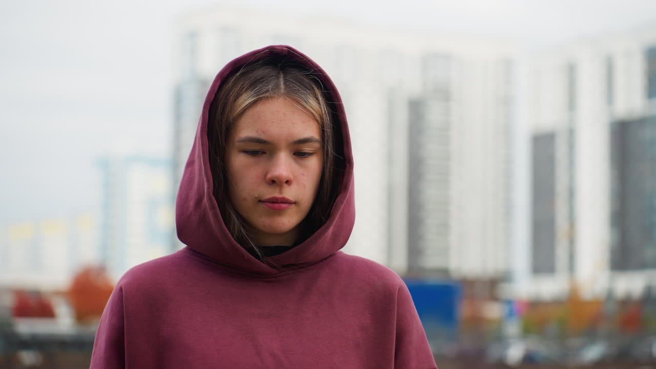 Workout participant in burgundy hoodie strolling urban walkway while glancing sideways at modern high rise buildings under cloudy sky, lined with spring flowers by garden and parked cars