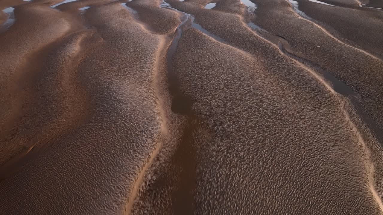 Fast flight over undulating sand patterns with seawater pools at sunset. Aerial footage.