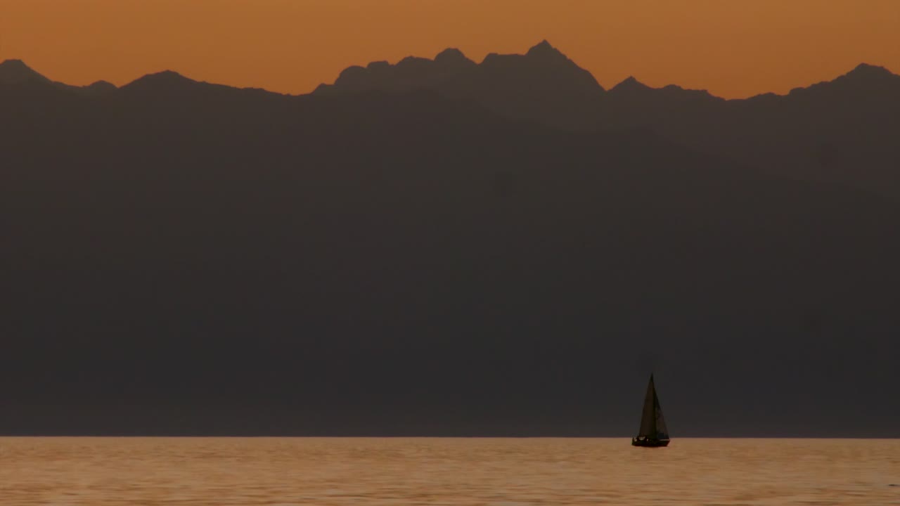 Optagelser af en stor sø ved solnedgang med en sejlbåd og et stort bjerg, der fremhæver rolig natur, smukke solnedgangsfarver, majestætiske bjerge, menneskelig aktivitet og naturlig kontrast