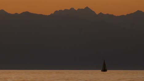 Footage-of-a-big-lake-at-sunset-with-a-sailing-boat-and-a-big-mountain,-showcases-serene-nature,-beautiful-sunset-colours,-majestic-mountains,-human-activity-and-natural-contrast