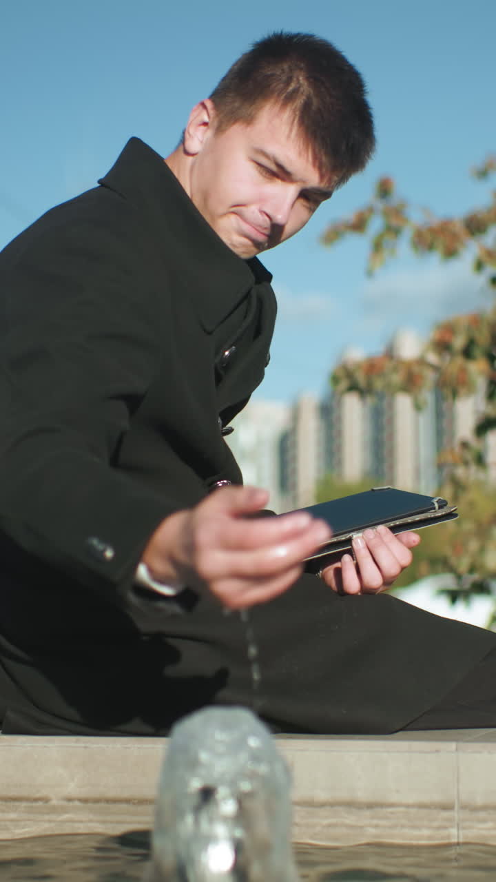 물 에 앉아있는 남자가 타블을 사용하면서 물 표면을 만지고 다리에 앉아 있으며, 도시 초록색, 현대적인 건물, 그리고 밝은 빛 하늘 아래 움직이는 자동차로 둘러싸여 있습니다.