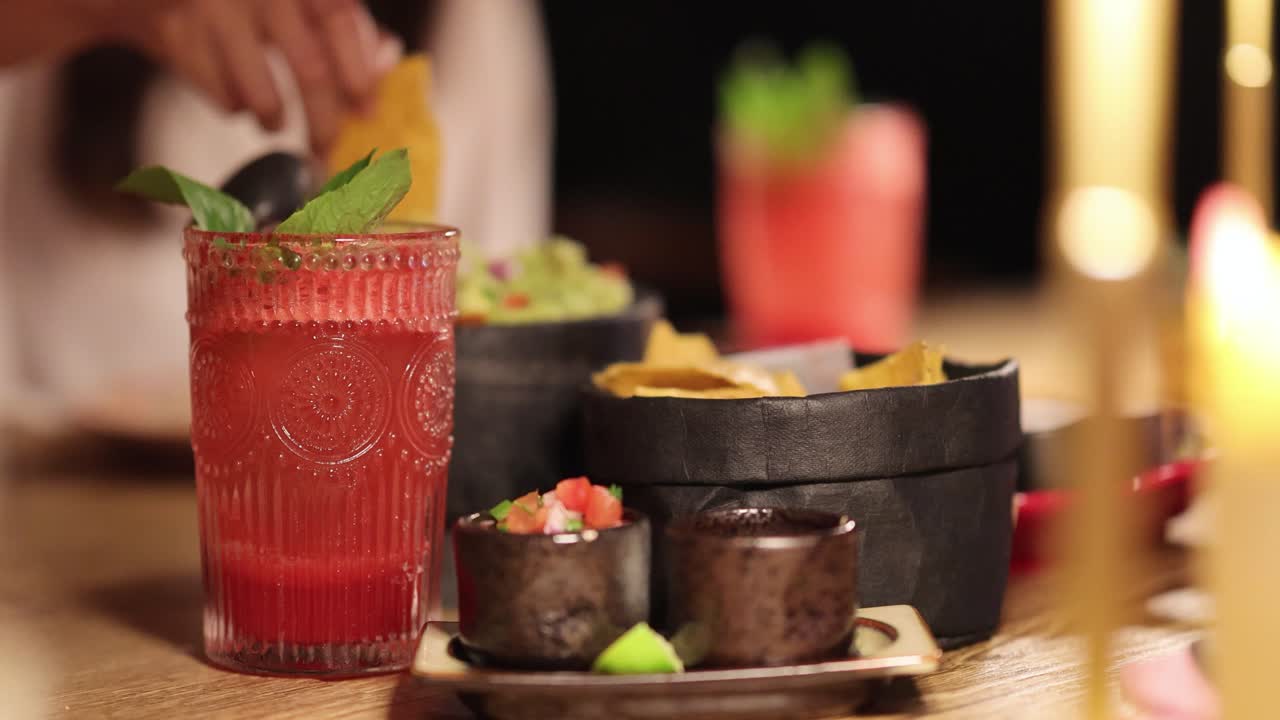 A hand dips chips into guacamole beside a vibrant drink, capturing a lively dining atmosphere with warm lighting