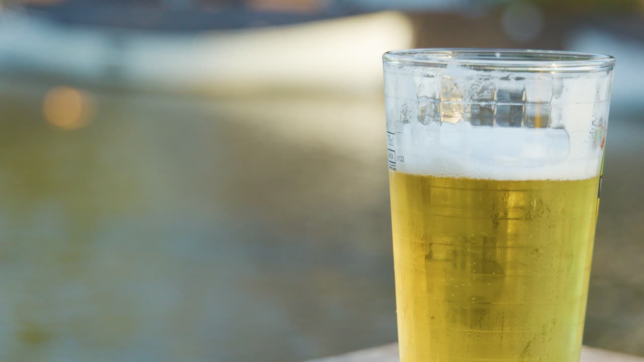 Hand sets down pint of pale ale on riverside table in daylight, blurred water background