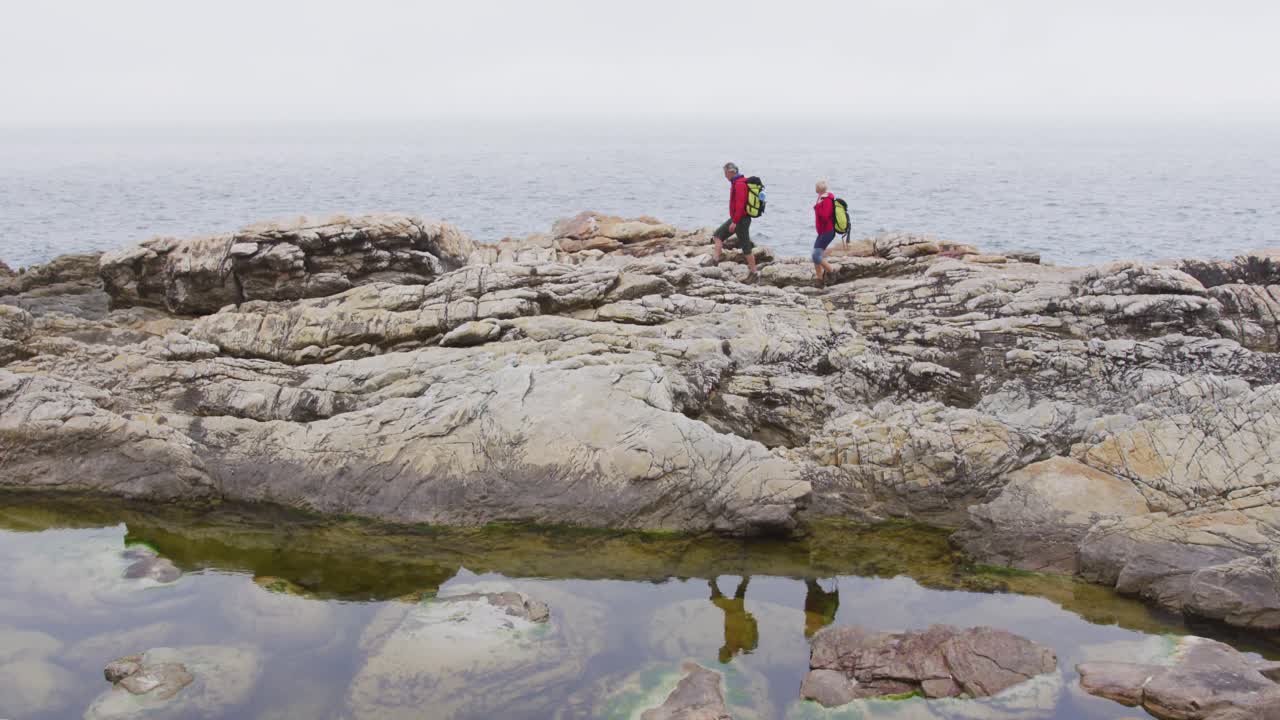 pareja de excursionistas mayores con mochilas caminando por las rocas mientras caminan cerca de la orilla del mar.