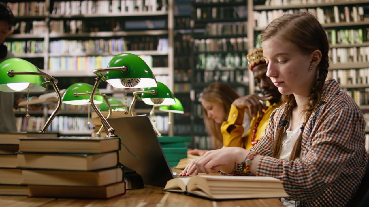 Giovane studentessa che lavora attorno a una scrivania della biblioteca con un laptop e una pila di libri
