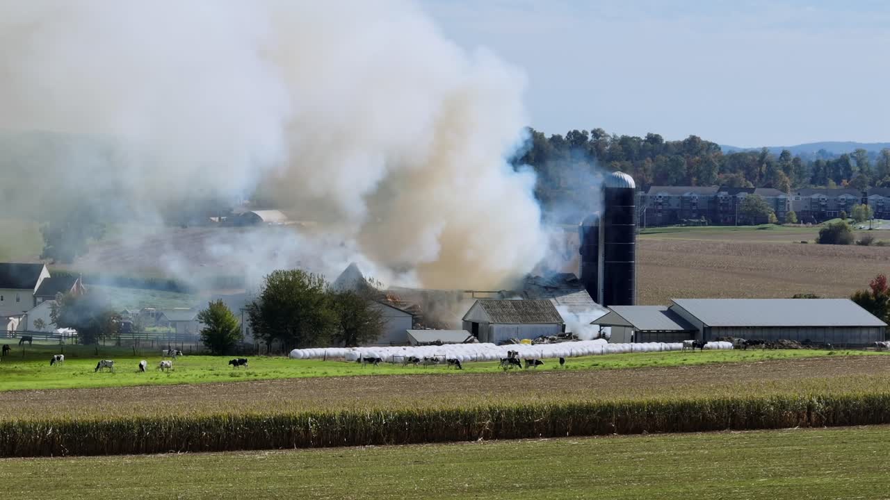 Aerial drone footage of a barn engulfed in flames on American countryside. Dark smoke drifting over rural fields and nearby houses, capturing dramatic fire and destruction at sunny day in USA. Wide
