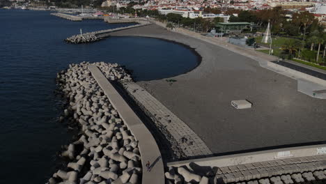 Toma-Aérea-Sobre-El-Muelle-Del-Puerto-De-La-Ciudad-De-Funcal-Y-Donde-Se-Puede-Ver-La-Playa-De-La-Ciudad-Así-Como-Las-Casas-Y-Edificios-De-La-Costa