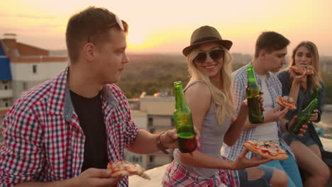 loving young couples sit on the roof and drink beer with pizza. they enjoy a beautiful sunset and time with their mates.