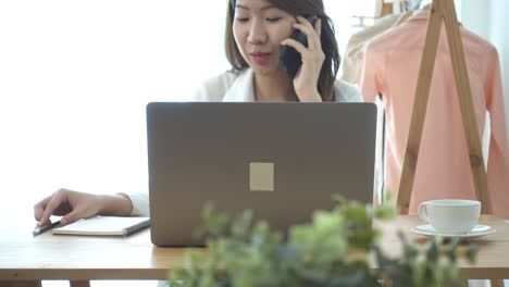 beautiful young smiling asian woman working on laptop while sitting in a living room at home. asian business woman using phone for work in her home office. enjoying time at home.