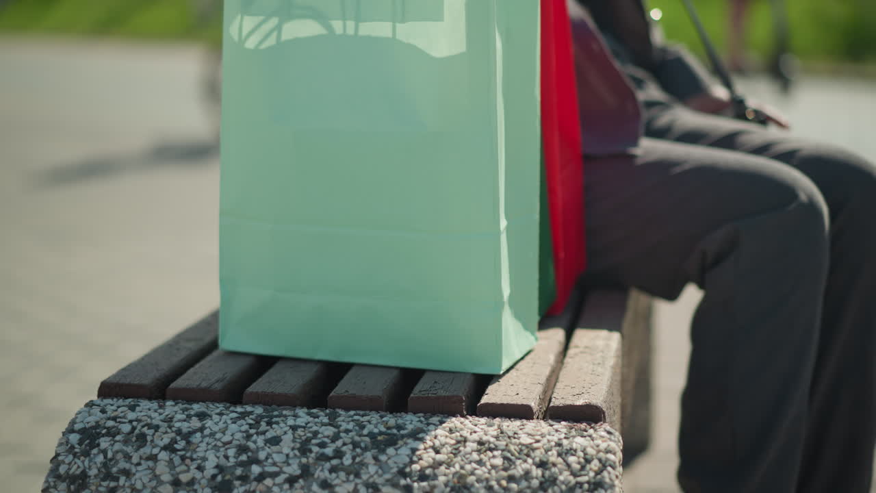 벤치에 쇼핑백을 놓고 앉아서 오른쪽 다리를 왼쪽 다리에 는 개인의 클로즈업 다리 시각, 자전거를 타는 사람을 특징으로하는 흐릿한 배경