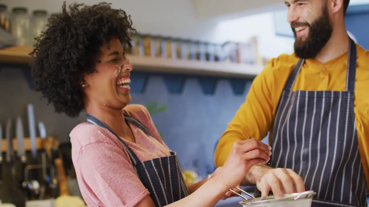 video de una pareja diversa riendo en delantales divirtiéndose y horneando juntos en la cocina en casa