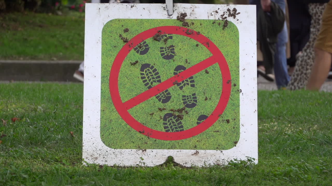 Close-up of a No Walking on Grass sign placed on a lawn with pedestrians walking in the background