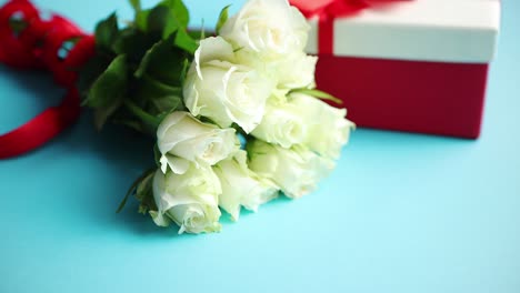 Strauß-Weißer-Rosen-Mit-Roter-Schleife-Auf-Blauem-Hintergrund-Verpacktes-Geschenk-An-Der-Seite