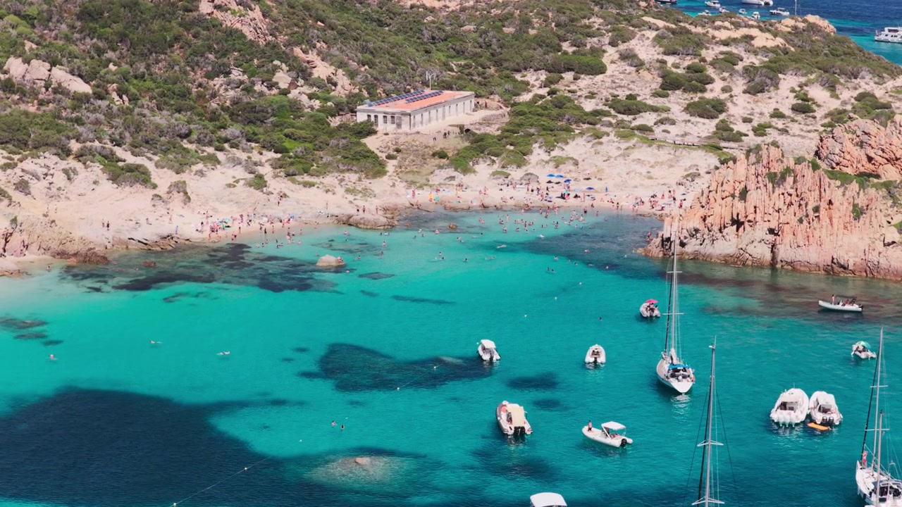 Vue aérienne en téléobjectif survolant des yachts à voile de luxe et des bateaux de tourisme, révélant une magnifique plage de sable nichée dans des falaises de granit sur l'île de Spargi dans l'archipel de la Maddalena en Sardaigne, Italie.