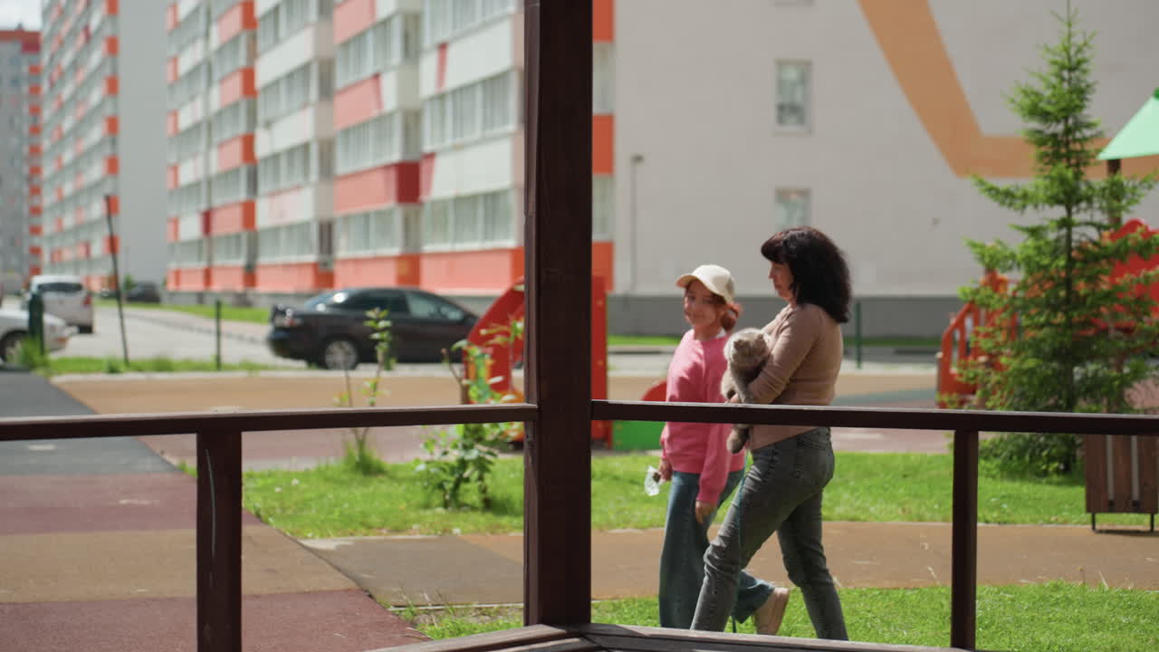 Women Calmly Pass By Park And City Elements, Two Women Stroll Silently Near Benches And Architectural Features, Two Women Quietly Walk Through Urban Setting Past Structures And Resting Benches