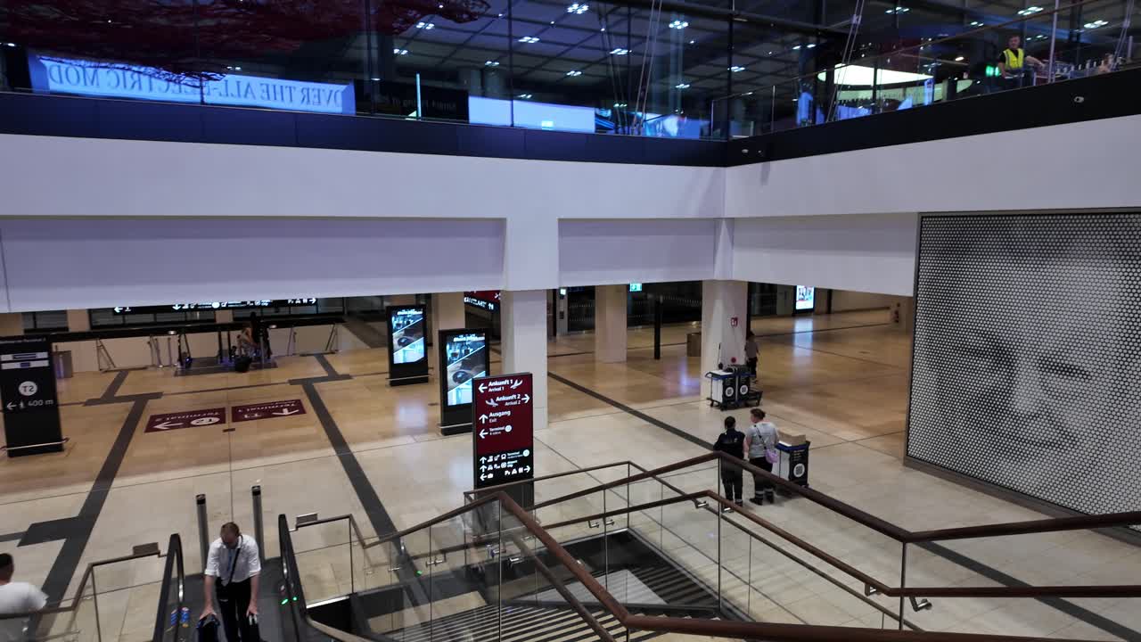 POV view of interior of Berlin Brandenburg Airport terminal with people using escalators and signage indicating directions to gates.