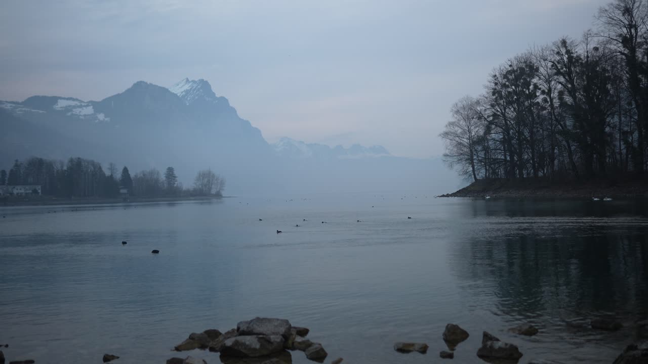 Timelapse of Lake Walensee, showcasing the serene beauty of the water reflecting mist-covered mountains, capturing the peaceful stillness and natural tranquility of the scene.