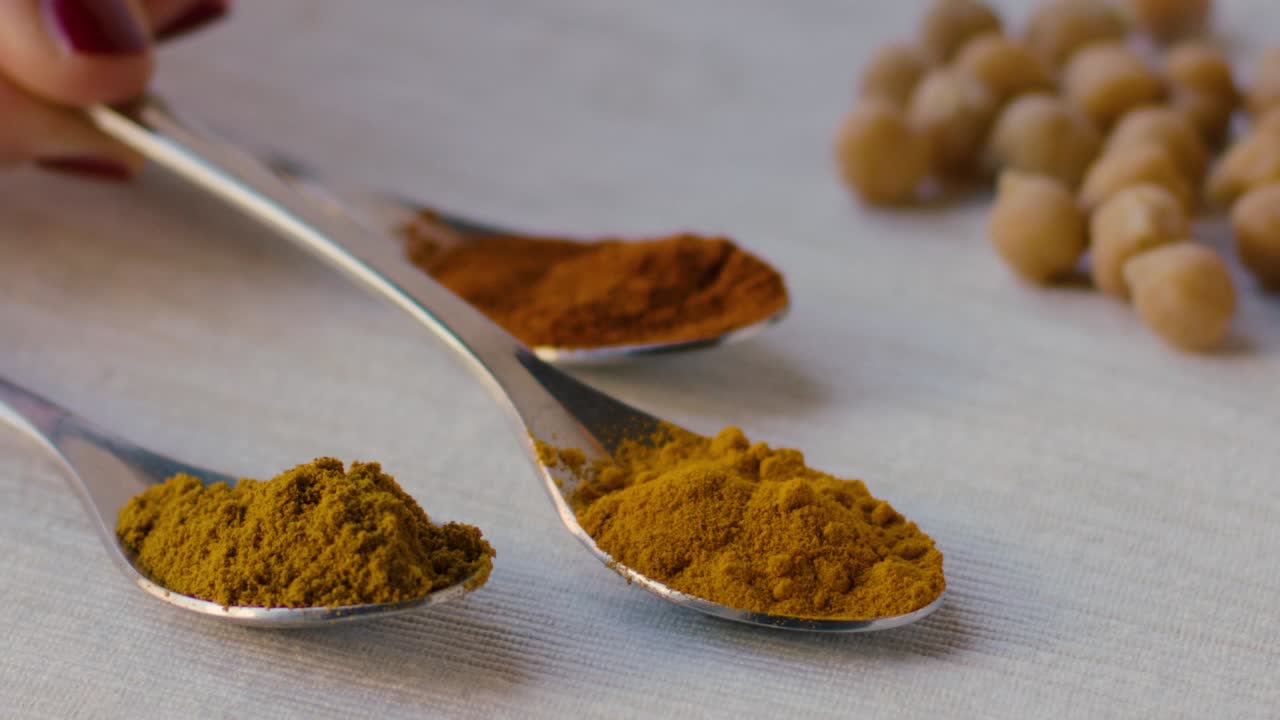 A spoonful of curry powder is placed on a table next to two other spoons with turmeric powder and saffron powder, chickpeas can be seen in the background